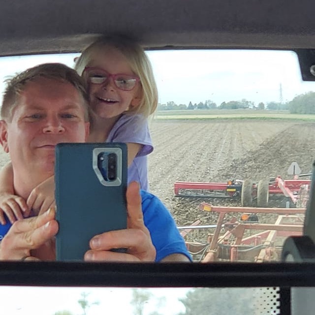A man taking a selfie of himself and his daughter in the rearview mirror of a tractor.