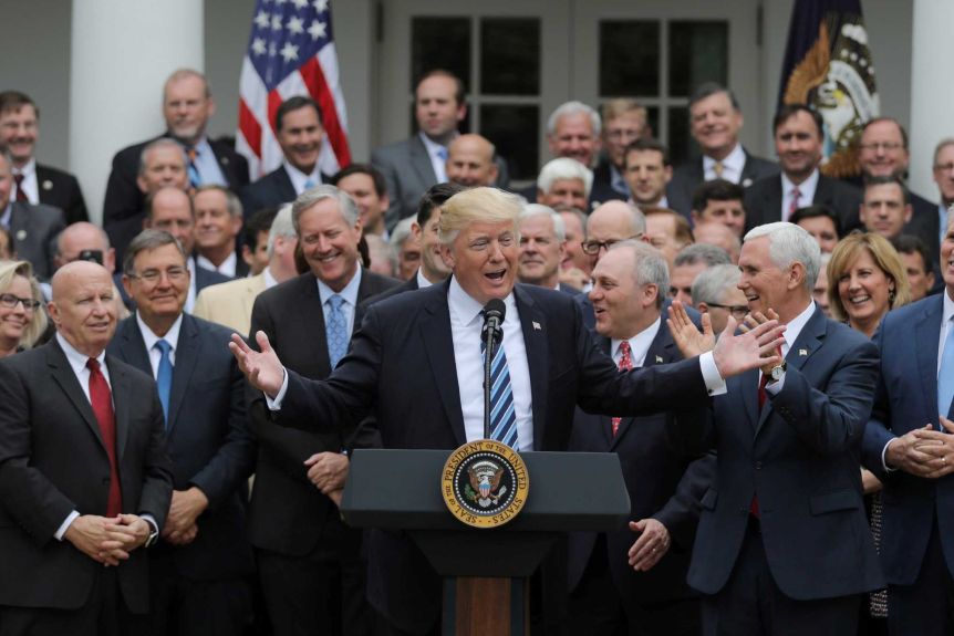 Donald Trump is behind a podium smiling while surrounded by smiling, clapping Republicans including Mark Meadows, Mike Pence and Steve Scalice.