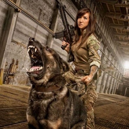 Female law enforcement officer in green fatigues holds a rifle and the leash of a barking police dog with its teeth displayed. 