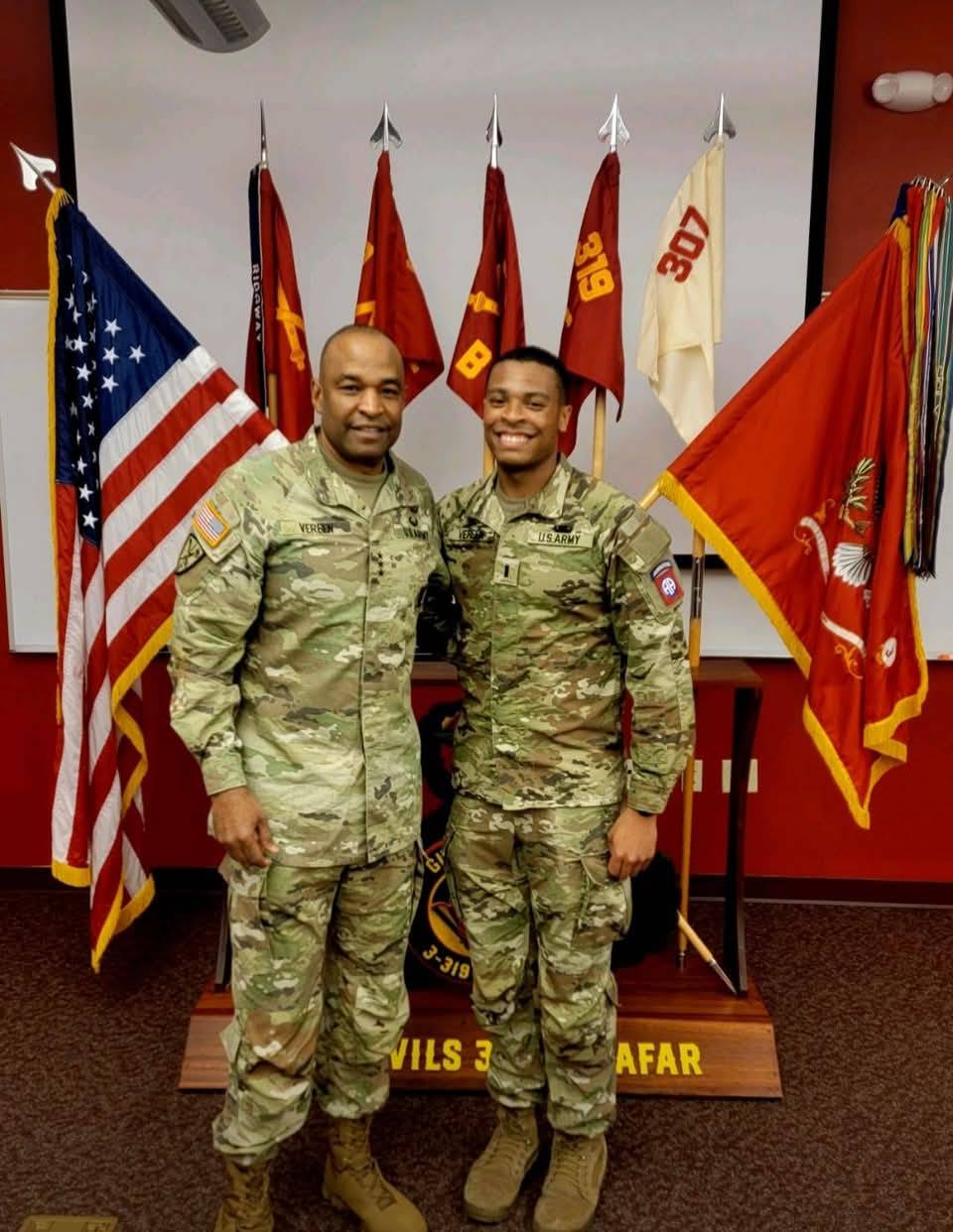 Two military personell posing in front of a stage with multiple flags.